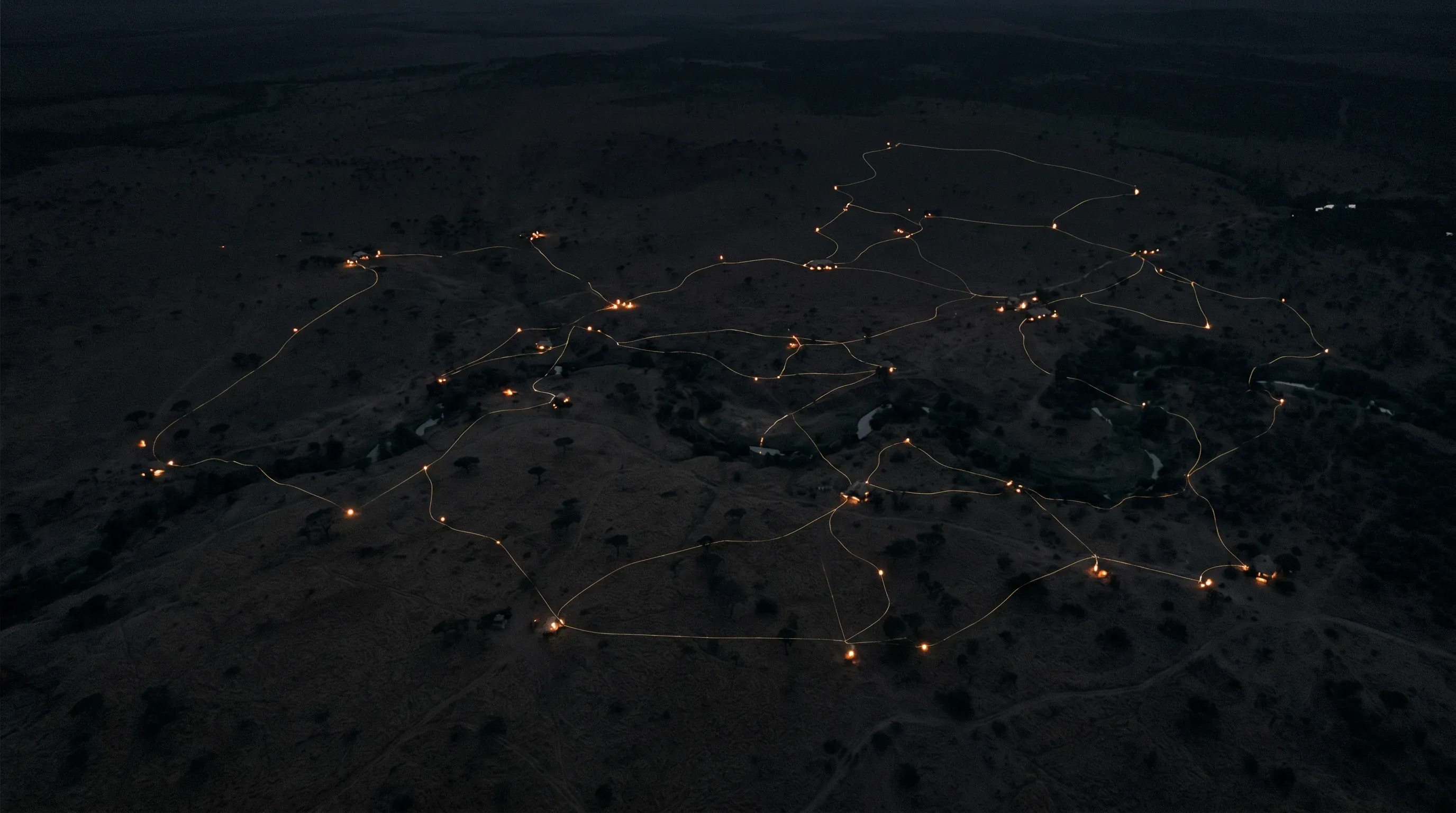 Night aerial of a wilderness: lit pathways link scattered camps into a single glowing network