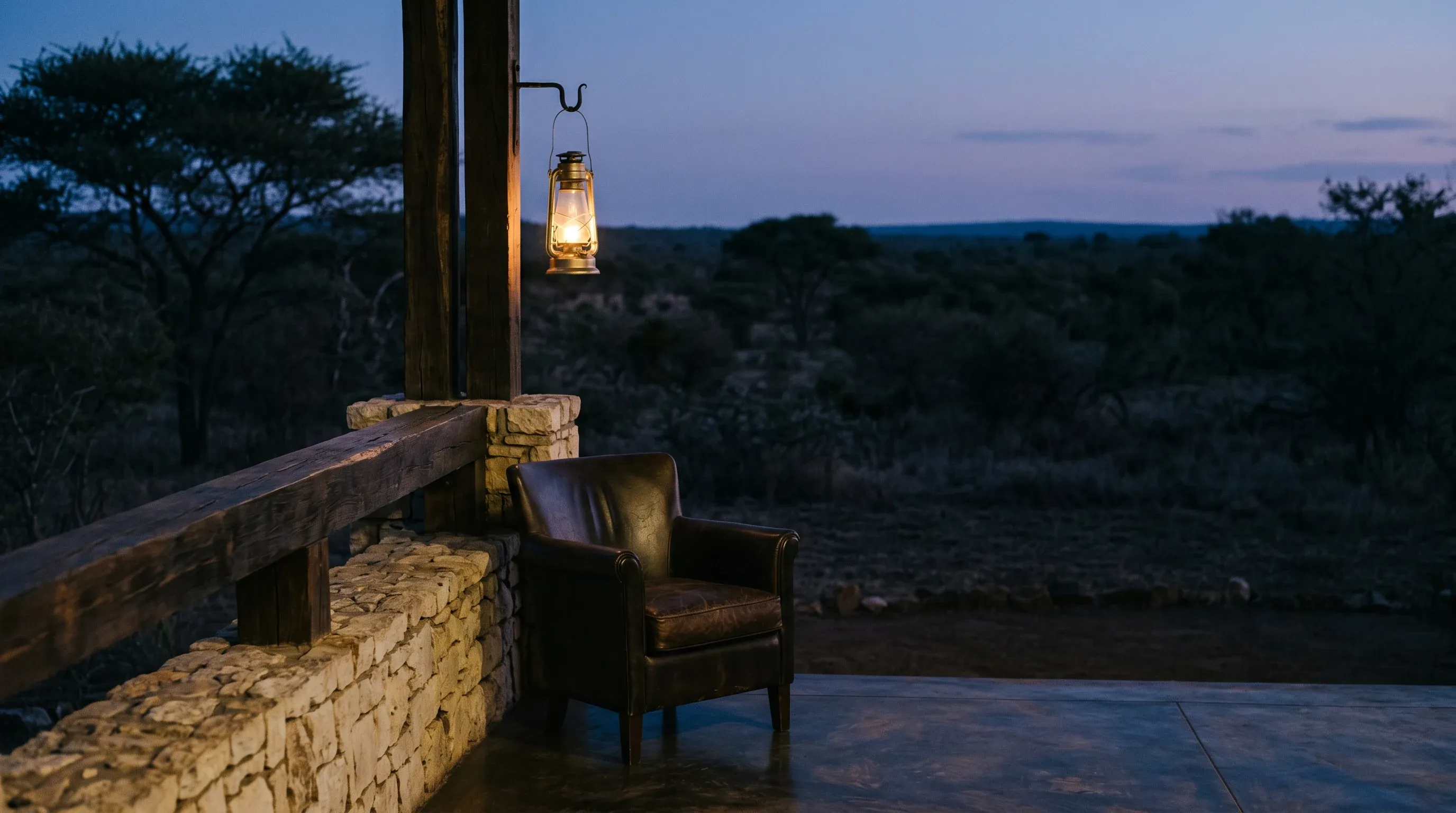 Lodge veranda at dusk: a lantern above a leather chair, stone wall and timber rail overlooking darkening bushveld
