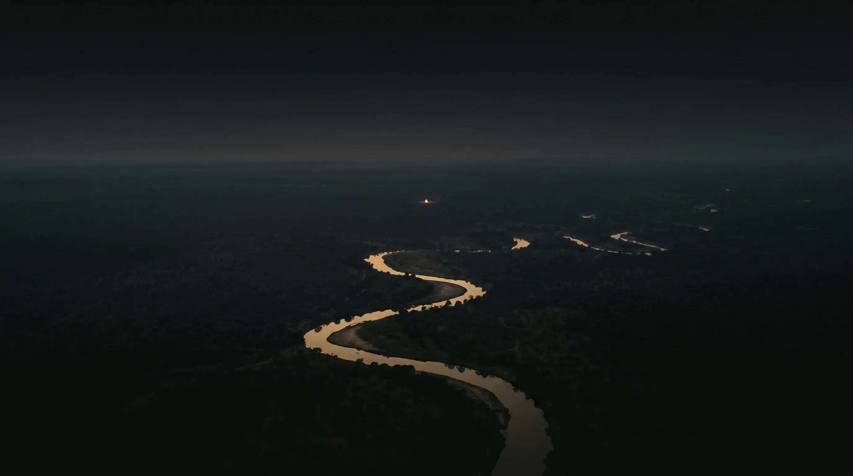 Aerial view of a river winding through dark wilderness at dusk, with a single lodge light at the water's edge